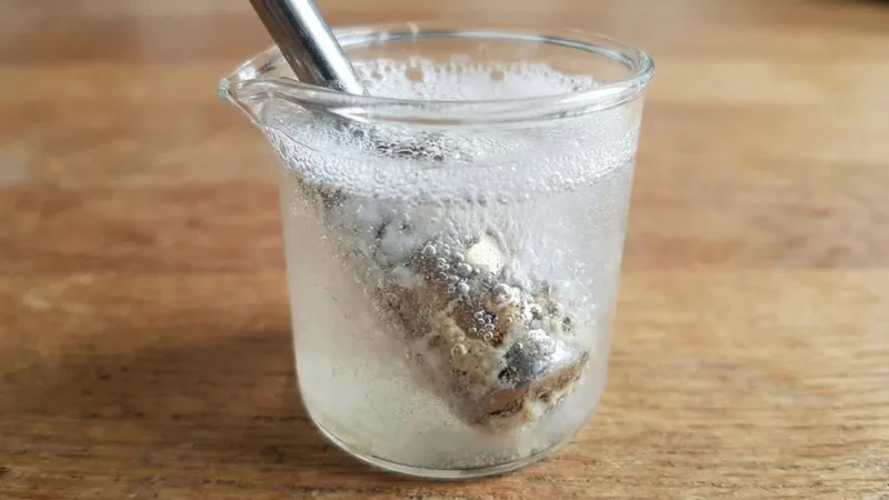 Steam wand tip submerged in a small glass container with cleaning solution, showing bubbles and the deep cleaning process
