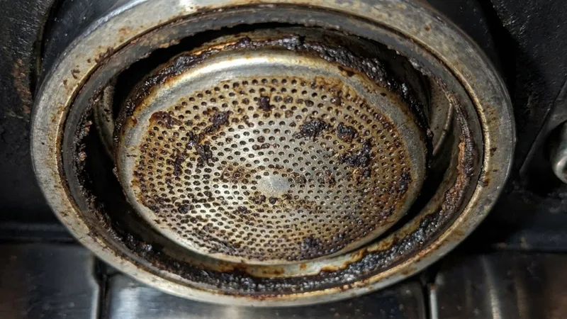 Extreme close-up of a heavily neglected shower screen still installed in the group head, showing dark brown coffee oil buildup, clogged holes, and crusty deposits around the edges
